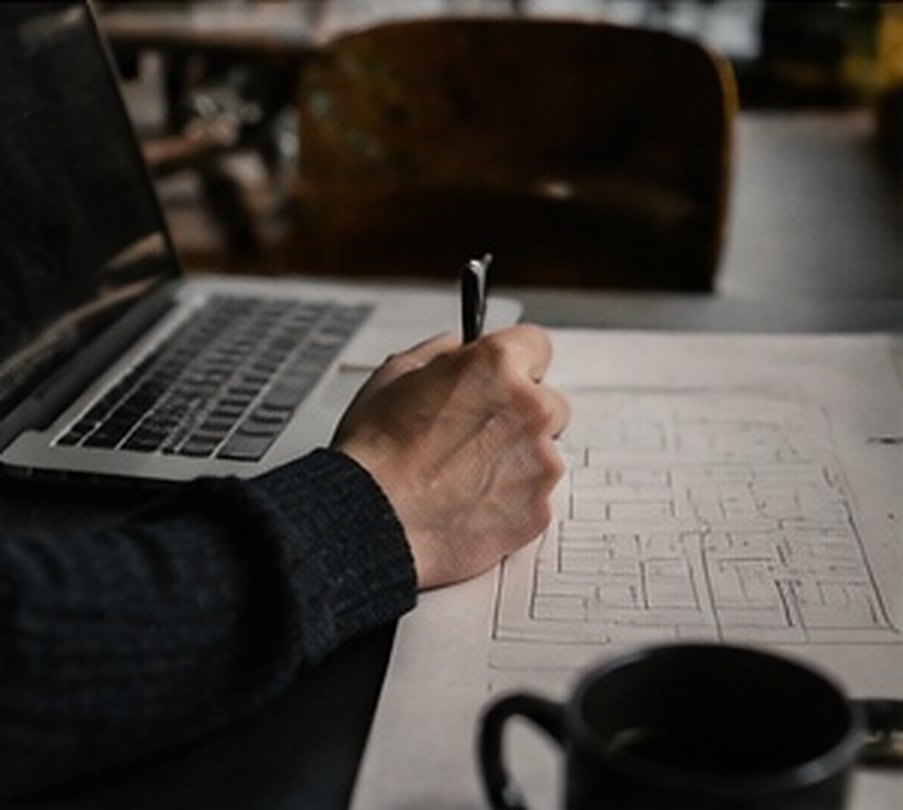 A hand drawing a plan in a notebook beside a cup of coffee